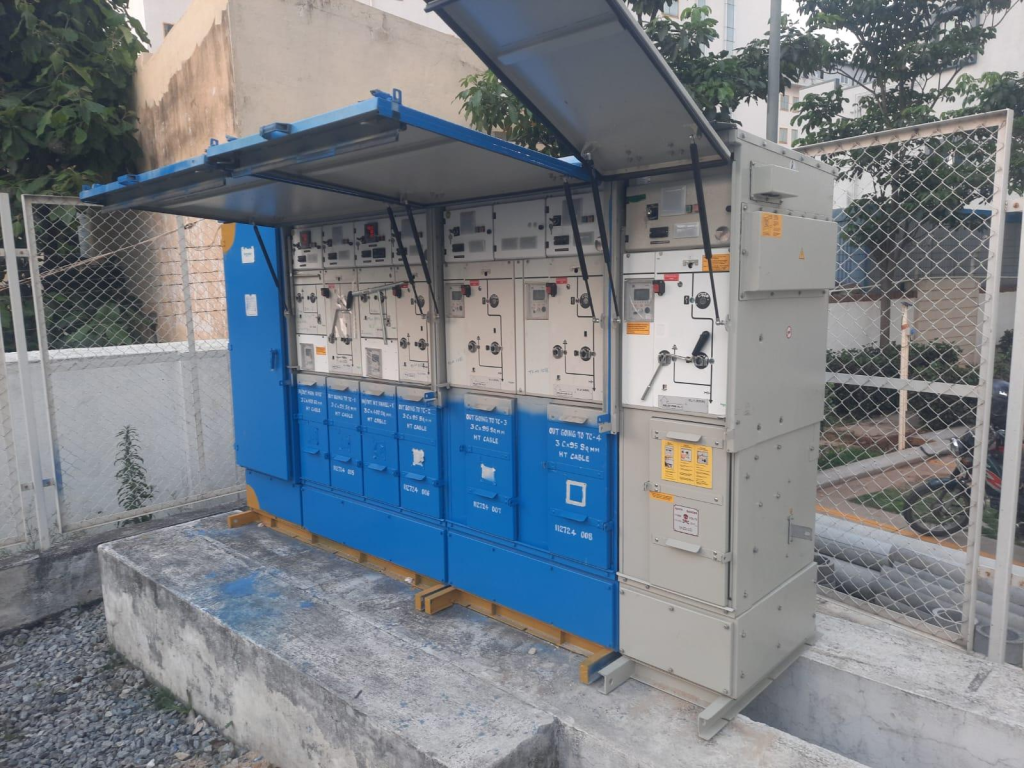 A modern urban power distribution network featuring Ring Main Unit equipment installed inside a compact substation or utility enclosure. The background includes commercial buildings, residential areas, and city streets, highlighting reliable electricity distribution for smart cities.